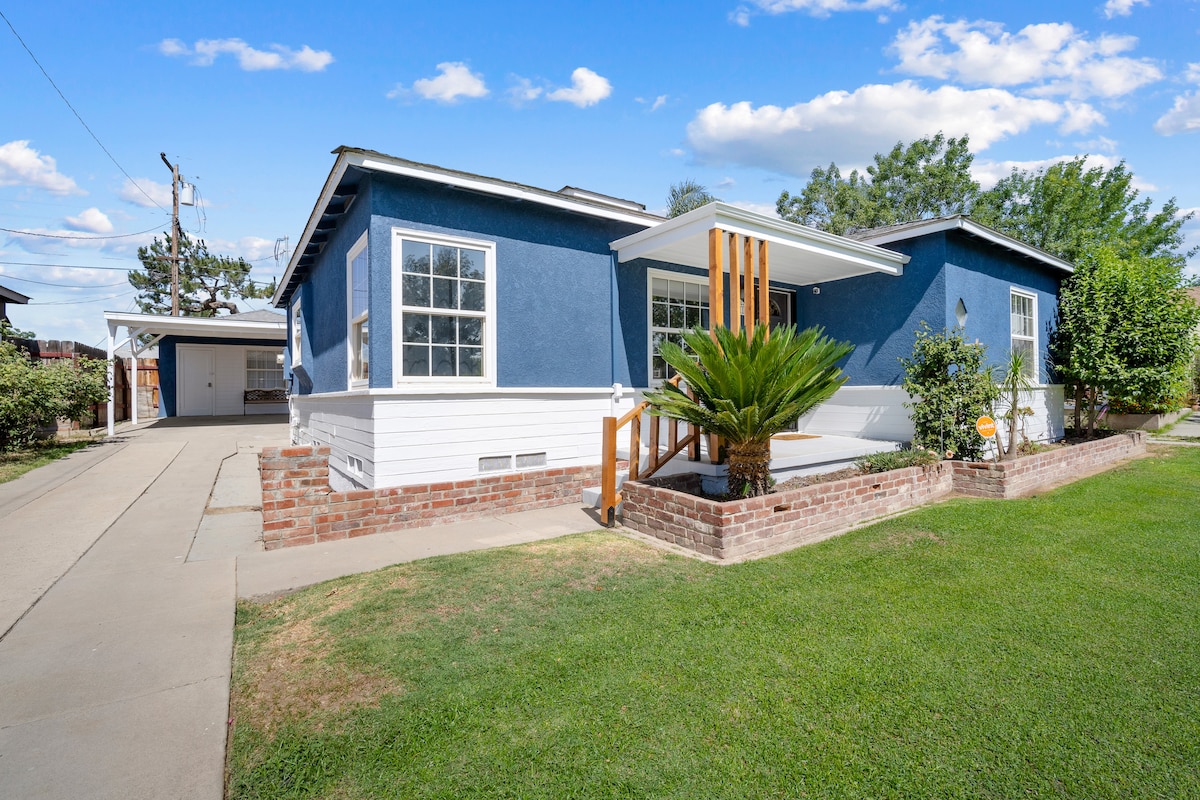 A single-story bungalow features a blue exterior with a white trim, surrounded by well-maintained grass. The entrance includes a small wooden deck with railing and a potted plant. The driveway leads to a garage, with trees and shrubs adding greenery beside the home.