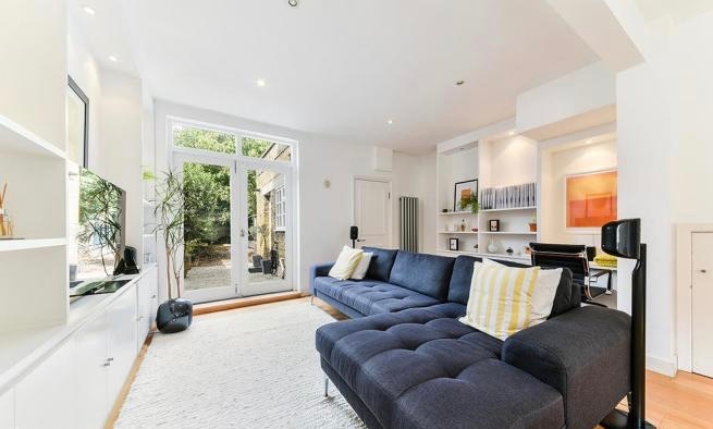 A modern living area features a spacious dark blue sectional sofa adorned with striped cushions. Natural light filters in through large glass doors that open to a private garden. A minimalist entertainment unit is positioned against the wall, accompanied by decorative shelving and plants.
