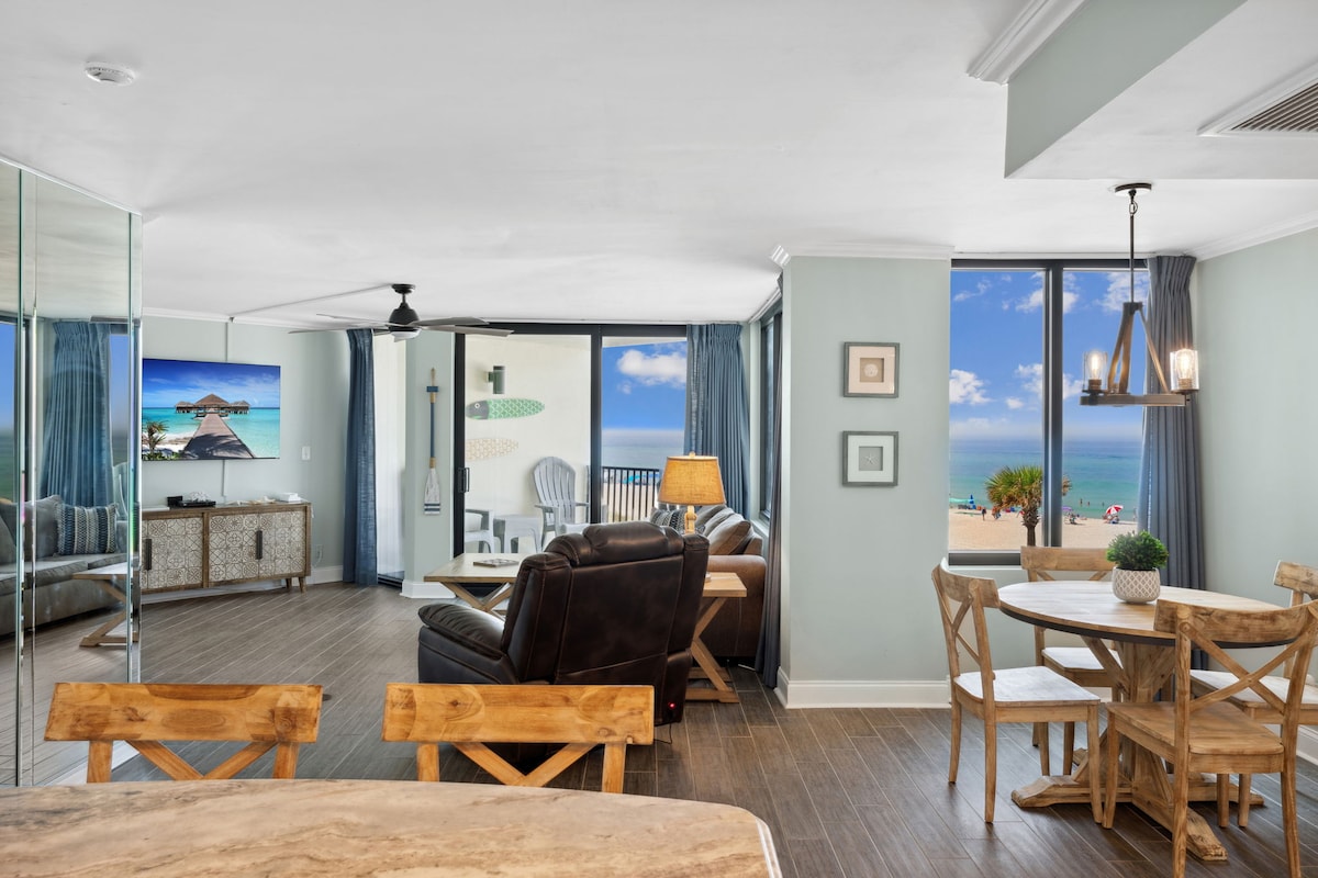 Sunlight is seen streaming through large windows, illuminating a spacious living area. Comfortable seating options, a dining table, and a view of the beach are featured. Decorative elements include a modern lamp and wall art, emphasizing a relaxing coastal atmosphere.