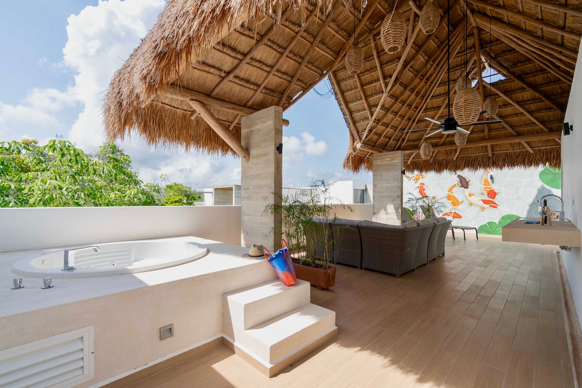 An open rooftop area features a thatched palapa covering, offering a private jacuzzi and comfortable seating. Steps lead down from the jacuzzi, and greenery is visible in the background. Brightly colored wall art adds a touch of vibrancy to the serene space.