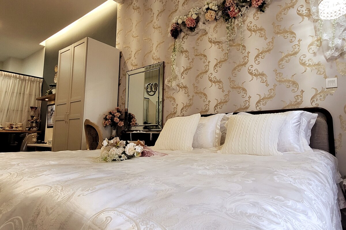 A spacious bed is adorned with elegant white bedding and decorative pillows. Floral accents are displayed on the wall above, complementing the intricate wallpaper design. A wardrobe and a mirror are positioned in the background, contributing to the room's stylish décor.