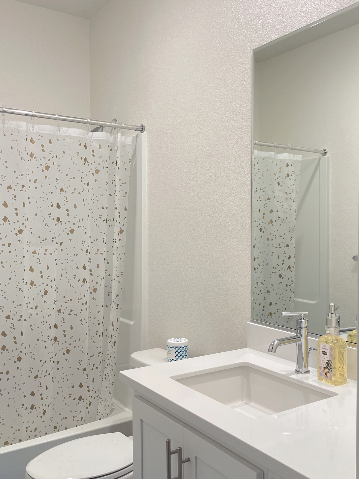 A modern bathroom features a clean design with a spacious sink and contemporary fixtures. A shower curtain with a gold speckled pattern provides a decorative touch. A large mirror reflects the bright environment, enhancing the overall functionality and style of the space.
