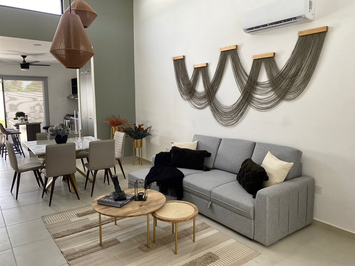 A spacious living area features a light grey sectional sofa complemented by dark cushions. Two round coffee tables are placed in front, with decorative items on top. The wall is adorned with a textured installation, and a dining area is visible in the background, highlighting a modern wooden table and elegant chairs.