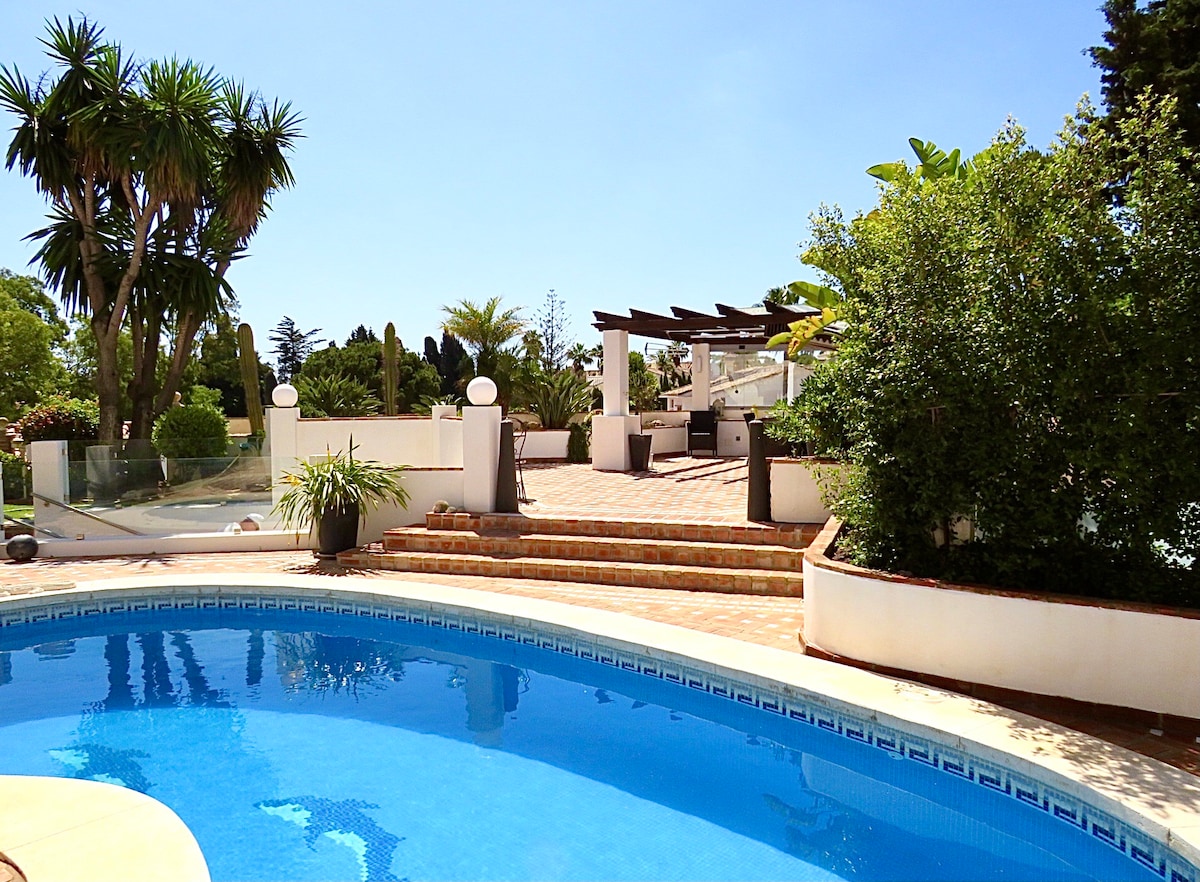 A private pool glistens under a clear blue sky, surrounded by lush tropical plants and elegant landscaping. Steps lead up to a spacious terrace outfitted with modern seating, providing a tranquil setting for relaxation amidst nature.