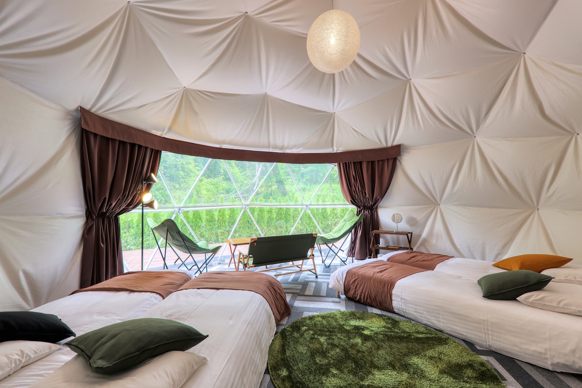 A spacious dome room features two single beds dressed in neutral tones, complemented by decorative pillows. A green area rug provides a cozy touch. Large windows offer panoramic views of the outdoors, while natural light brightens the interior, accented by warm curtains and a contemporary light fixture.