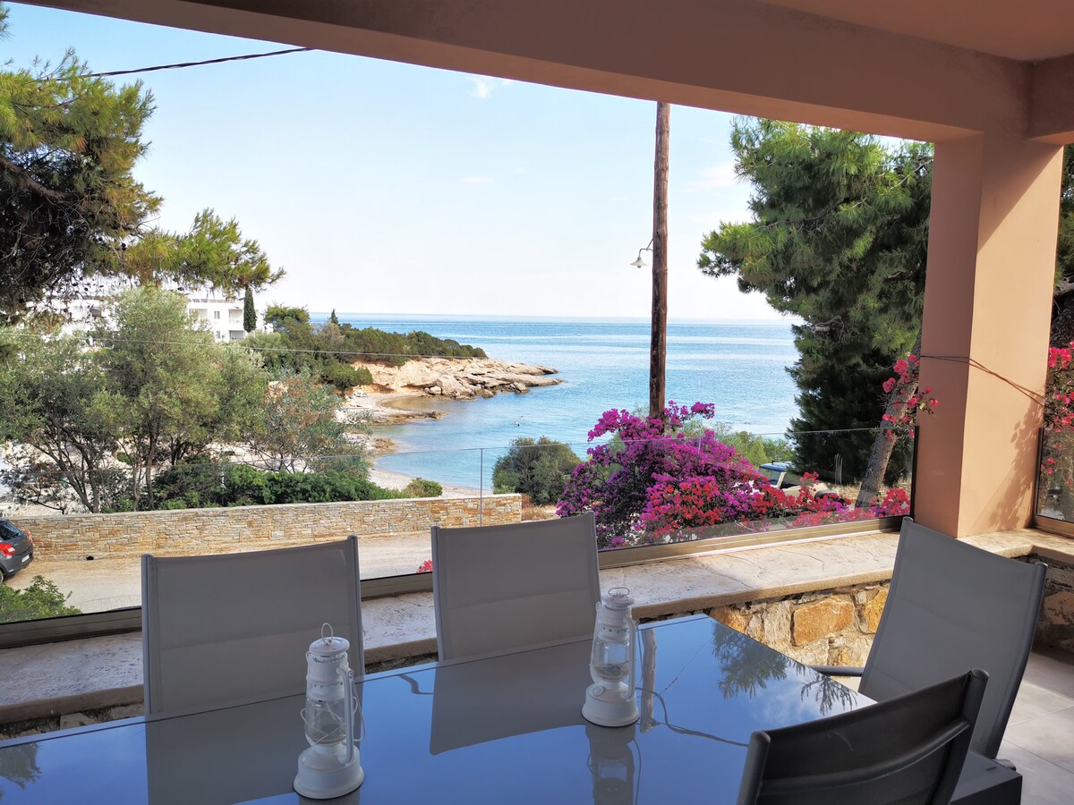 A spacious outdoor dining area features a glass table surrounded by several white chairs. A view of the calm sea and rocky shoreline is visible in the background, framed by vibrant flowering plants and greenery.