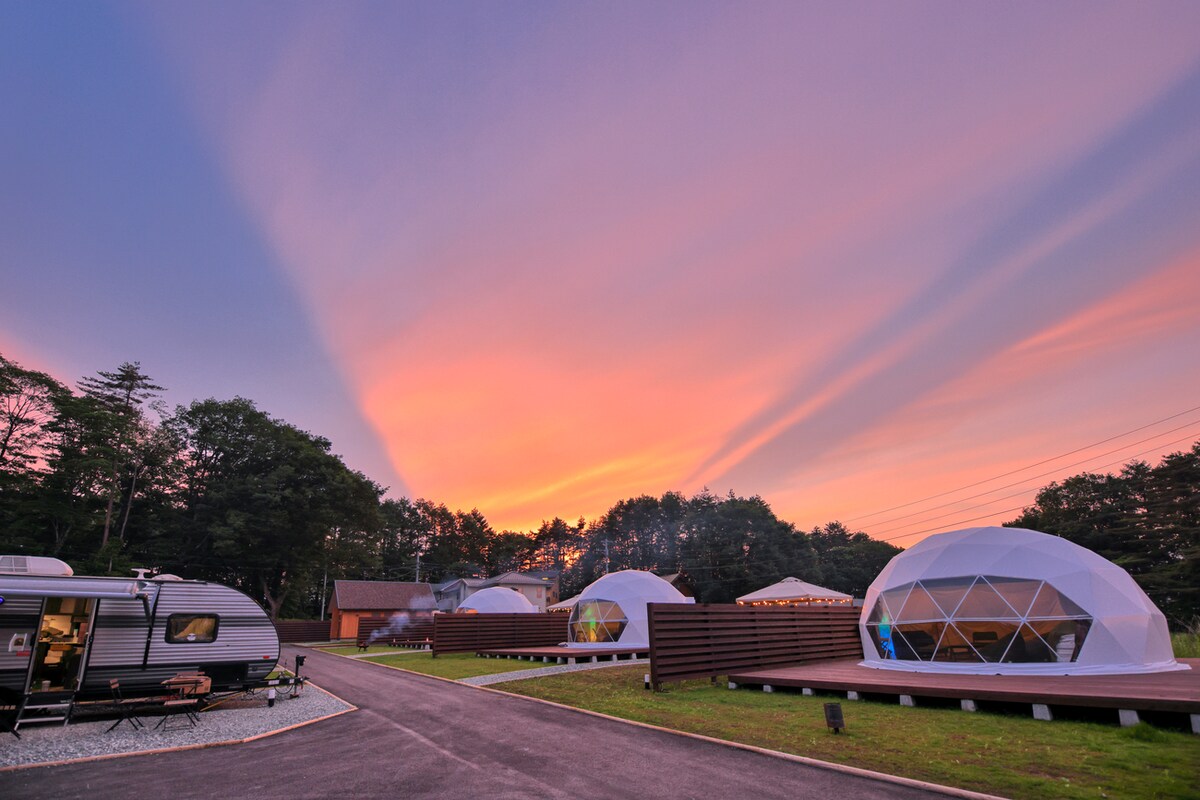Dome tents are positioned in a spacious outdoor area, surrounded by lush greenery. A pastel sky at sunset adds a warm glow, while a caravan is parked nearby. The geometric shapes of the domes contrast with the natural landscape, inviting exploration and adventure.