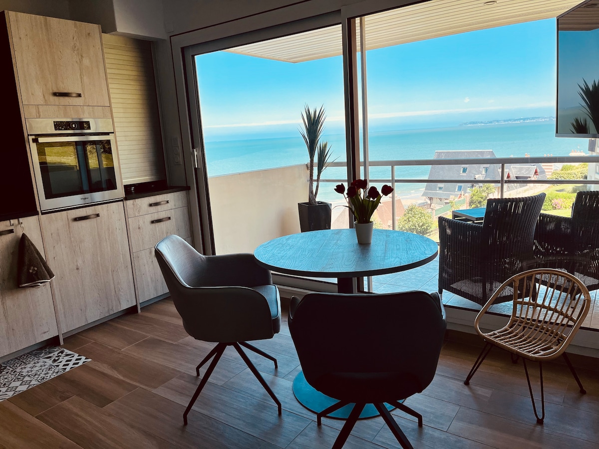 A bright dining area features a round table surrounded by modern seating options. Large glass doors offer a panoramic view of the ocean, enhancing the space with natural light. Potted plants add a touch of greenery, complementing the coastal scenery outside.