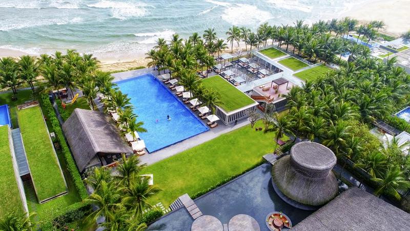 An aerial view captures a resort with multiple pools set amidst lush greenery. A large infinity pool extends toward the beach, surrounded by palm trees and sun loungers. The shoreline and waves are visible beyond the pool area, contributing to the serene atmosphere.