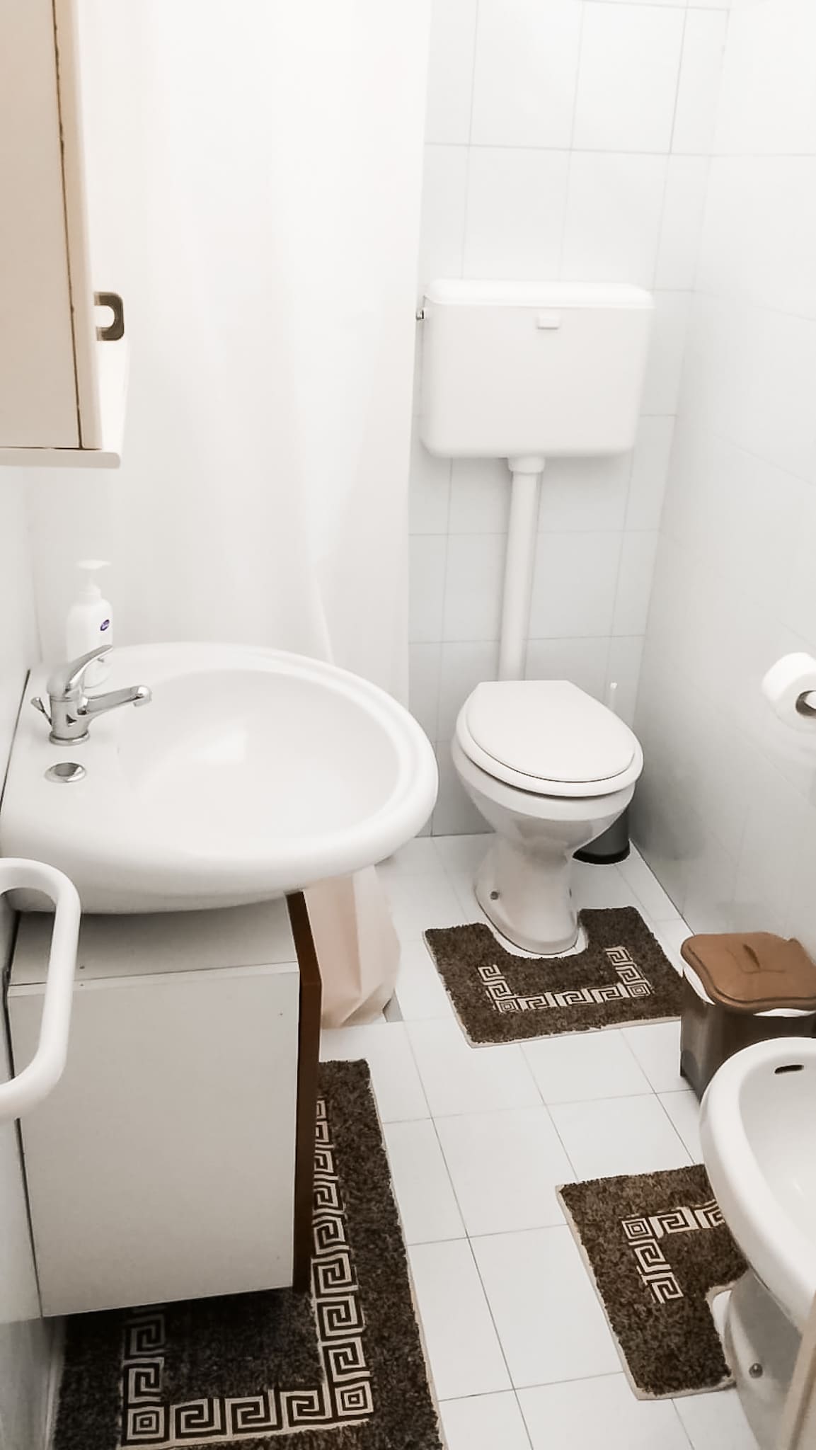 A clean bathroom space is presented with white tiles and light-colored fixtures. A round sink is set on a cabinet, accompanied by a toilet. Two small rugs add a touch of warmth to the floor. A shower curtain offers privacy and separation.