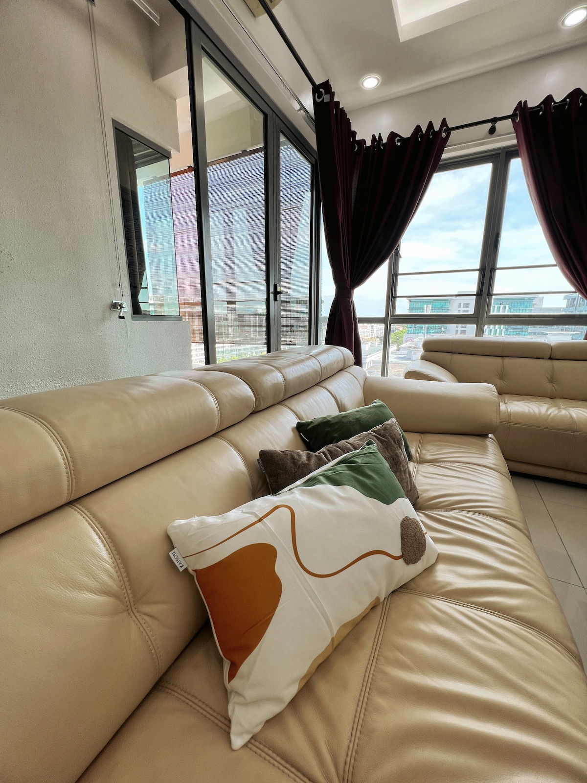 A light-colored leather sofa is positioned next to expansive windows, allowing natural light to fill the space. Toss pillows with abstract patterns are arranged on the sofa, complemented by rich burgundy curtains framing the view of the cityscape.