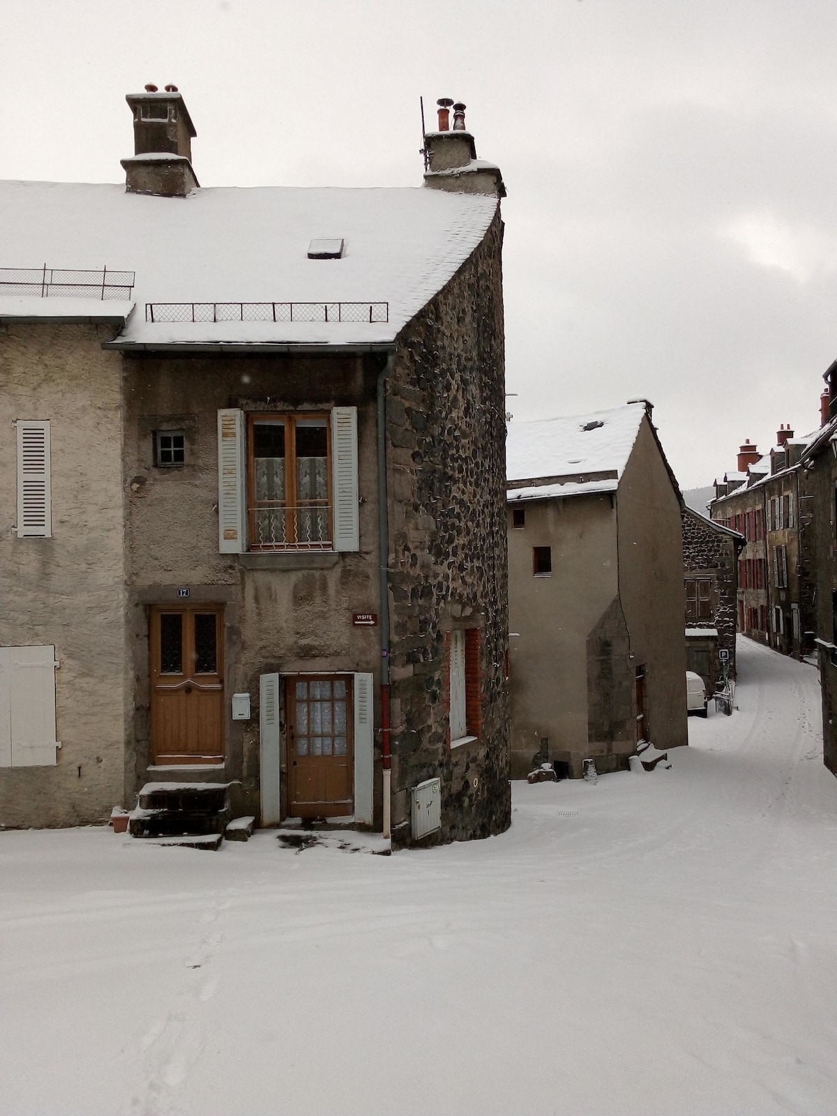 A snow-covered street winds through a quaint historic village, showcasing the unique architecture of stone and weathered facades. The two-story building features a wooden door, framed windows, and snow resting gently on the rooftops, creating a serene winter scene.