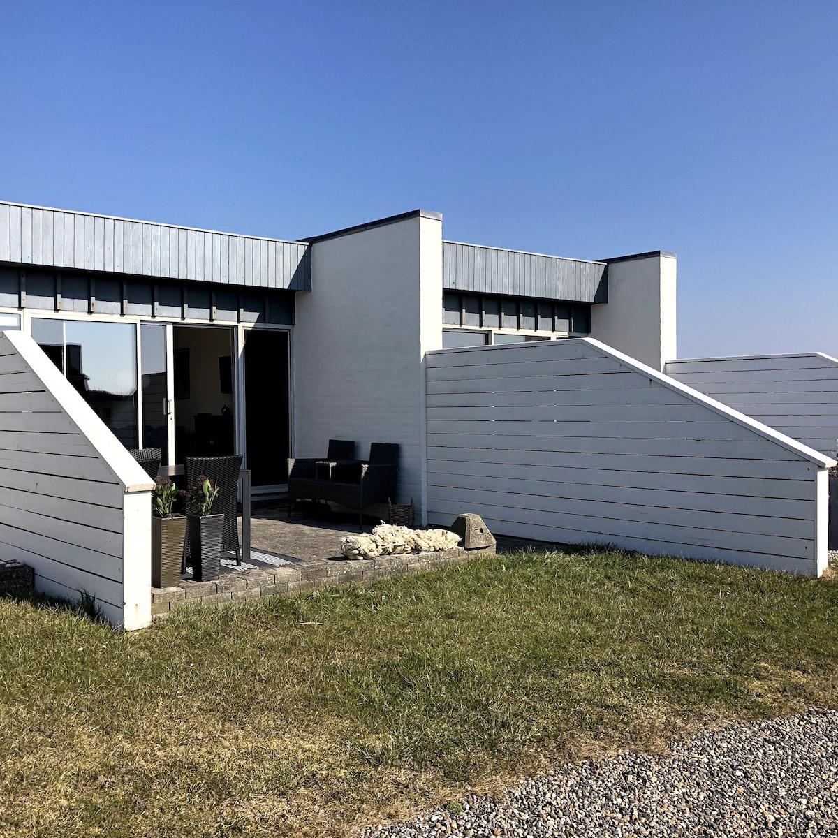 The exterior of the newly renovated holiday home is depicted, showcasing clean lines and a modern design. A patio area is visible, featuring outdoor furniture. The surroundings include a grassy area and a clear blue sky, creating an airy and open atmosphere.