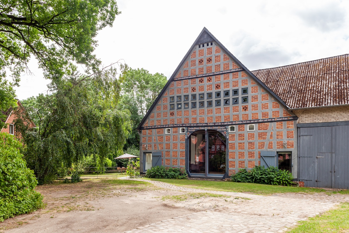The exterior of a traditional barn-style building is shown, featuring a distinctive façade with a mix of brick and wooden elements. Large arched windows provide views into the interior. Surrounding greenery and outdoor seating areas create a natural ambiance.