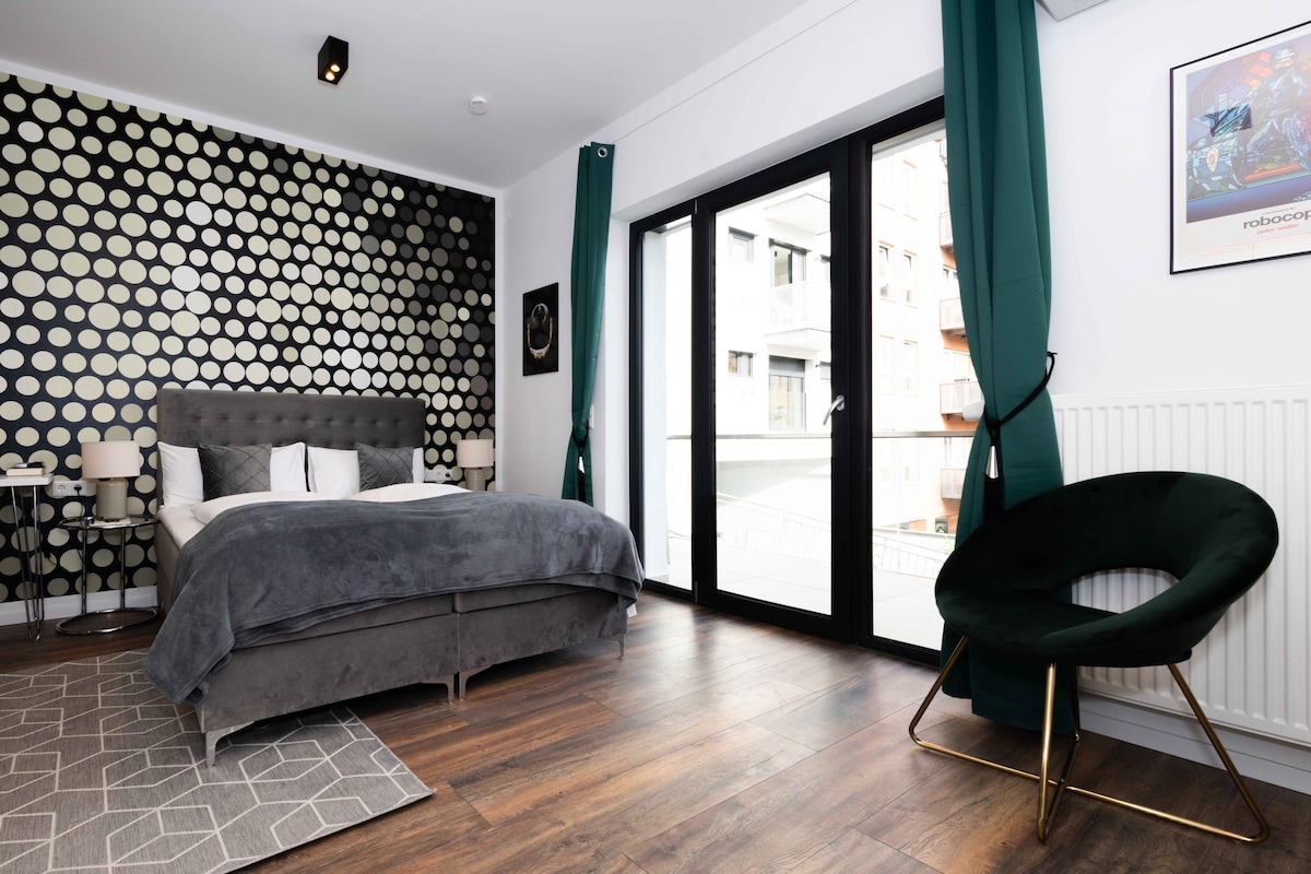 A stylish bedroom features a large bed with gray bedding against a modern dotted accent wall. Floor-to-ceiling glass doors allow natural light to flood in and provide access to a balcony. A circular green chair complements the contemporary decor.