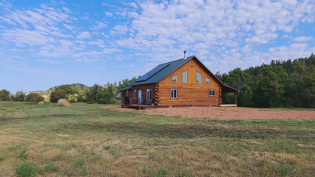 A spacious log cabin is seen set against a backdrop of clear blue skies and scattered clouds. The building features multiple large windows and a generous porch, while surrounding grassy fields are punctuated by round hay bales and greenery.