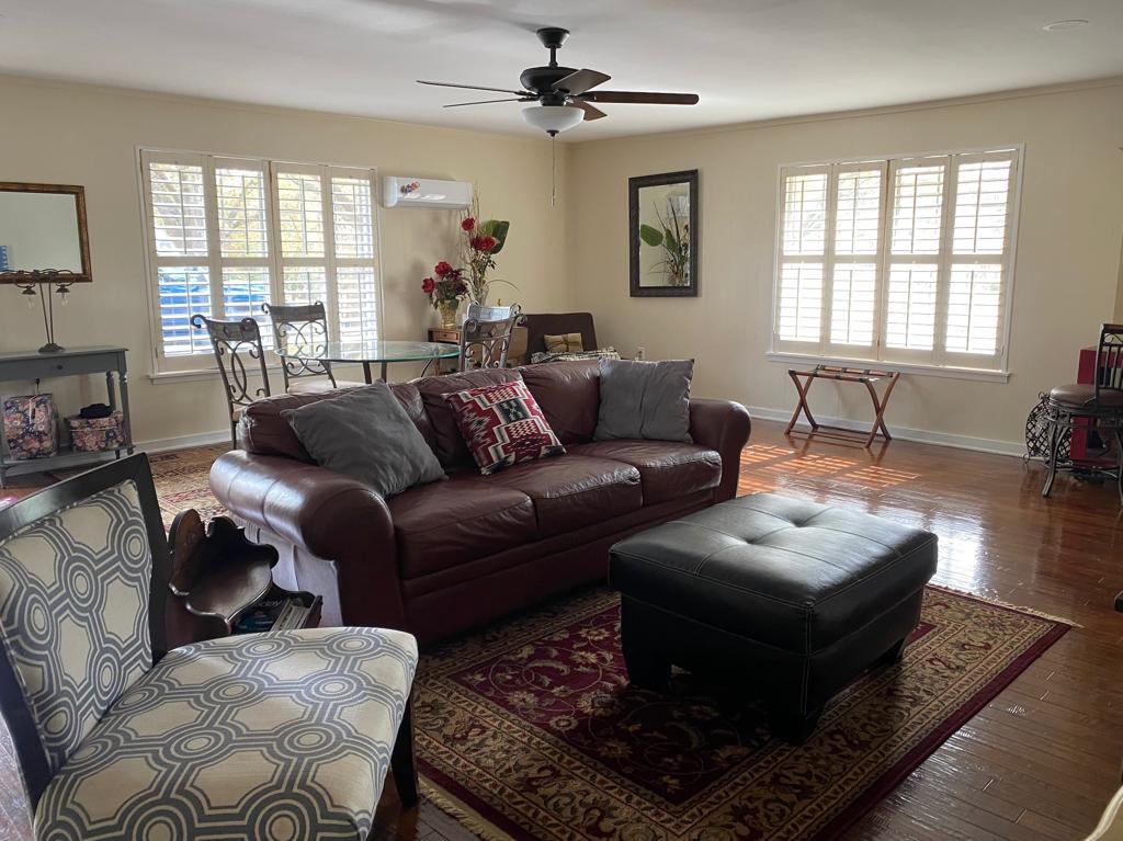 The living area features a comfortable leather sofa and an ottoman, complemented by a patterned accent chair. Natural light enters through multiple windows with wooden shutters, illuminating the space. A dining table and chairs are visible in the background, along with decorative elements.