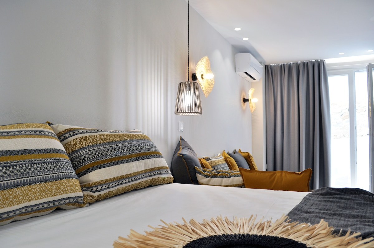 A well-appointed bedroom features a spacious bed adorned with textured pillows. Soft lighting is provided by hanging fixtures, while a seating area with accent cushions enhances the comfort. Large windows invite natural light, complemented by neutral walls and warm textiles.