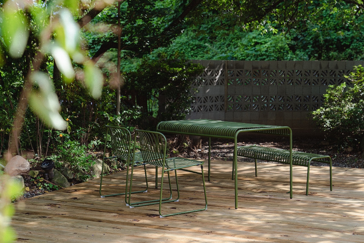 An outdoor seating area is surrounded by greenery, featuring a rectangular table and two chairs, all in a matching green metal design. The decking has a natural wood finish, providing a harmonious connection with the garden environment.