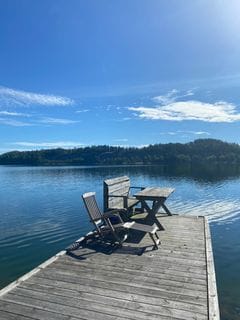 A wooden dock extends over a calm body of water, featuring a table and two chairs positioned for relaxation. Lush green hills are visible in the background under a clear blue sky, reflecting softly on the water's surface.