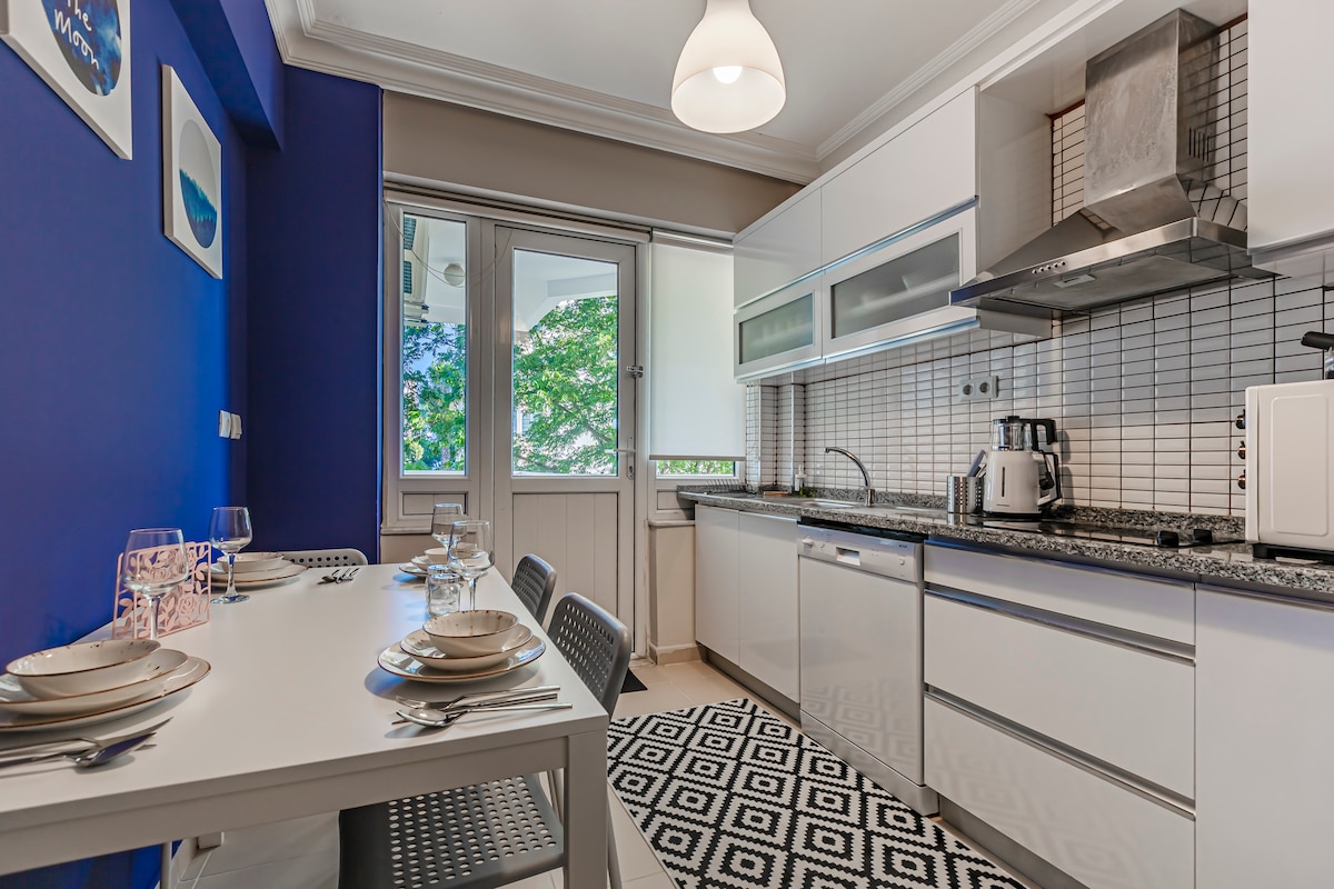 A modern kitchen features sleek white cabinetry and a black granite countertop. A dining table is set for four, and a patterned rug adds visual interest. Natural light enters through large windows, providing a view of the greenery outside, enhancing the space's brightness.