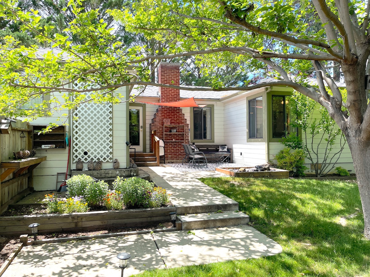 The exterior of a cottage is shown, featuring a well-maintained lawn with green grass and flower beds. A pathway leads to a deck with seating options, and a brick chimney adds character to the home. Trees provide shade in the inviting outdoor space.