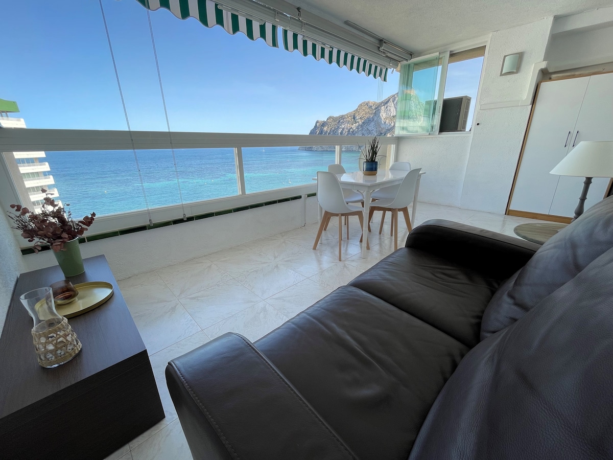 A spacious living area is visible, featuring a comfortable dark leather sofa and a modern dining set for four. Large glass doors offer a view of the sea and nearby cliffs, allowing natural light to flood the space. A small decorative table stands beside the sofa.