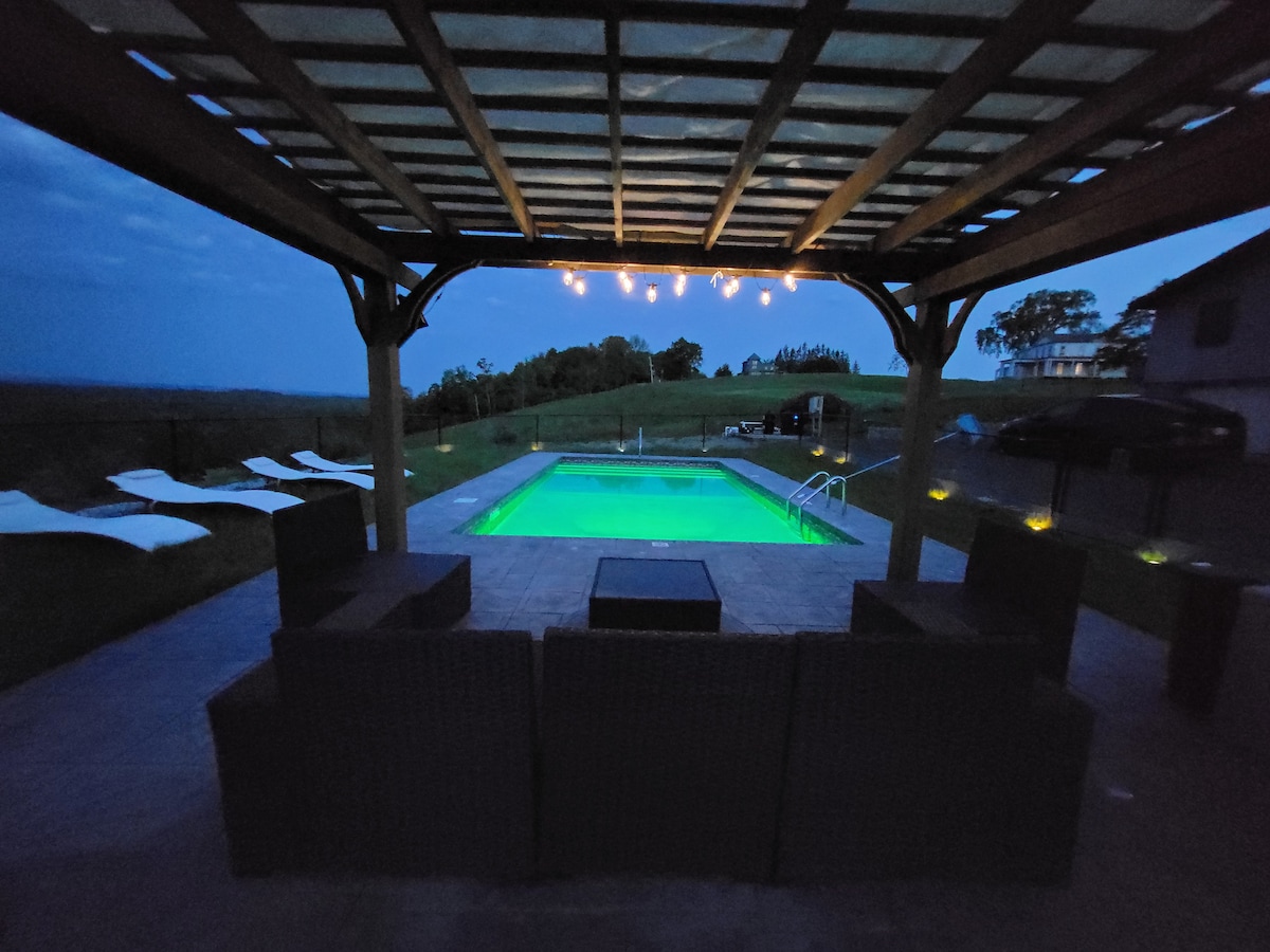 An evening scene reveals a spacious pool area beneath a wooden pergola. The illuminated pool glows in vibrant green, surrounded by stylish outdoor seating. Lounge chairs are positioned along the poolside, with gentle lighting enhancing the tranquil atmosphere.