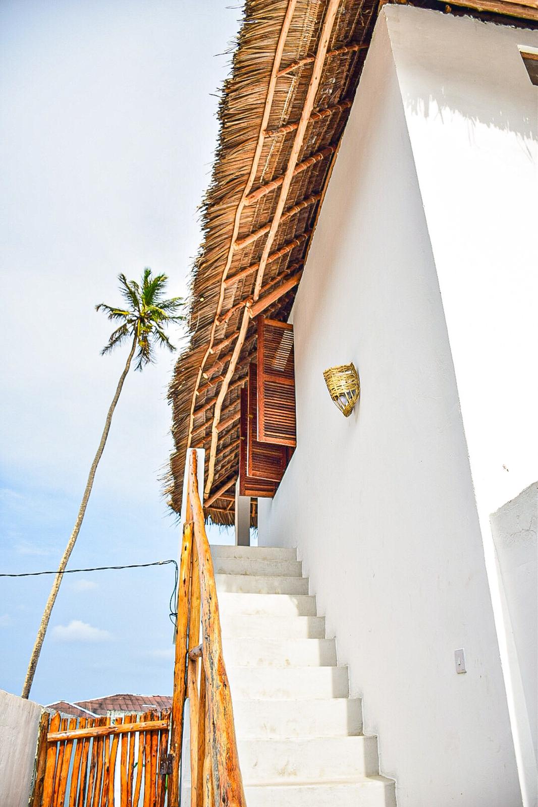 The exterior of a beach house is depicted, with a thatched roof and white walls. A set of concrete stairs leads to a wooden door, partially opened. A palm tree stands tall in the background under a clear sky.