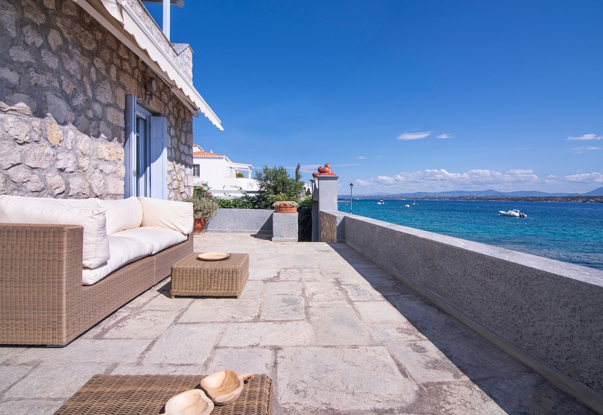 A spacious outdoor seating area is shown with a comfortable wicker sofa and a low coffee table on a stone patio. The surrounding landscape includes views of the clear blue sea and distant boats, with a sunny sky and soft clouds enhancing the scene.