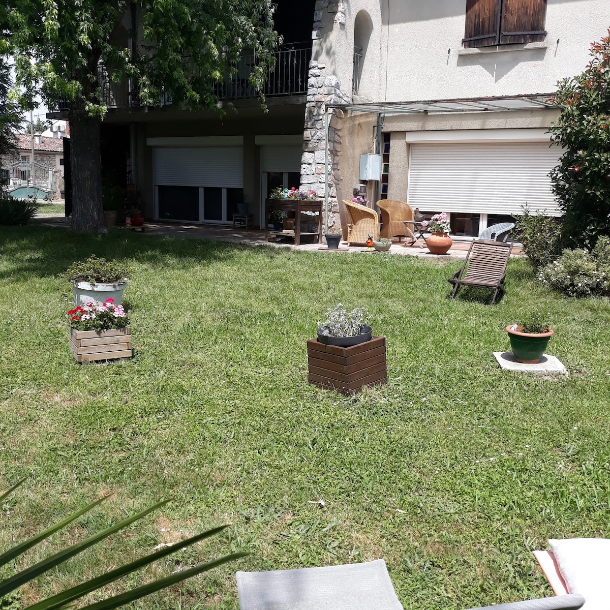 A well-maintained garden area features green grass, planters with blooming flowers, and a wooden lounge chair. The exterior of a two-story building is visible, showcasing stone accents and large windows. The space offers an inviting spot for relaxation amidst nature.