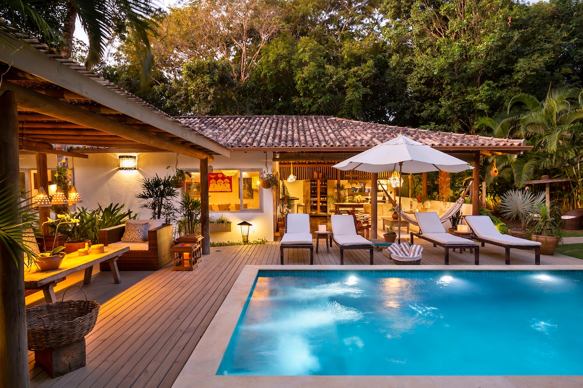 A serene outdoor living area is presented with a crystal-clear pool surrounded by loungers. An umbrella provides shade over the seating arrangement, while lush greenery frames the space. The charming architecture features a thatched roof, blending seamlessly with the tropical surroundings.