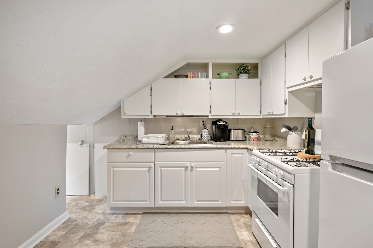 The kitchen area features modern cabinets and a countertop, providing ample space for meal preparation. A gas stove, refrigerator, and coffee maker are present, along with essential dishware and utensils. The sloped ceiling adds character to this well-lit, functional space.