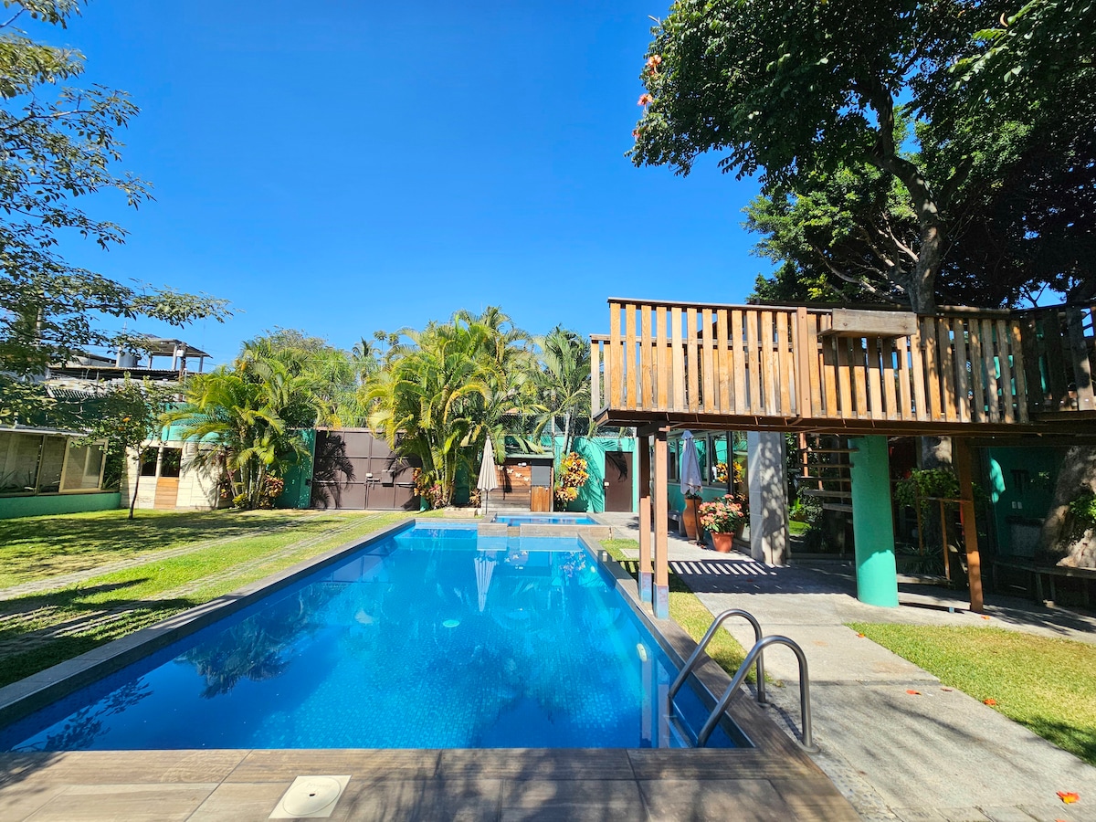 The image captures a clear blue pool surrounded by lush greenery. A wooden structure is visible in the background, along with various plants and trees that enhance the garden atmosphere. Bright sunlight illuminates the area, emphasizing the inviting space.