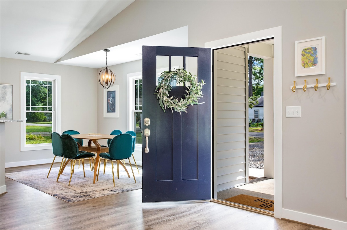A modern entryway is highlighted by a dark blue front door, adorned with a green wreath. The open space features a dining area with a circular wooden table surrounded by upholstered chairs. Large windows allow natural light to fill the room, emphasizing a welcoming atmosphere.