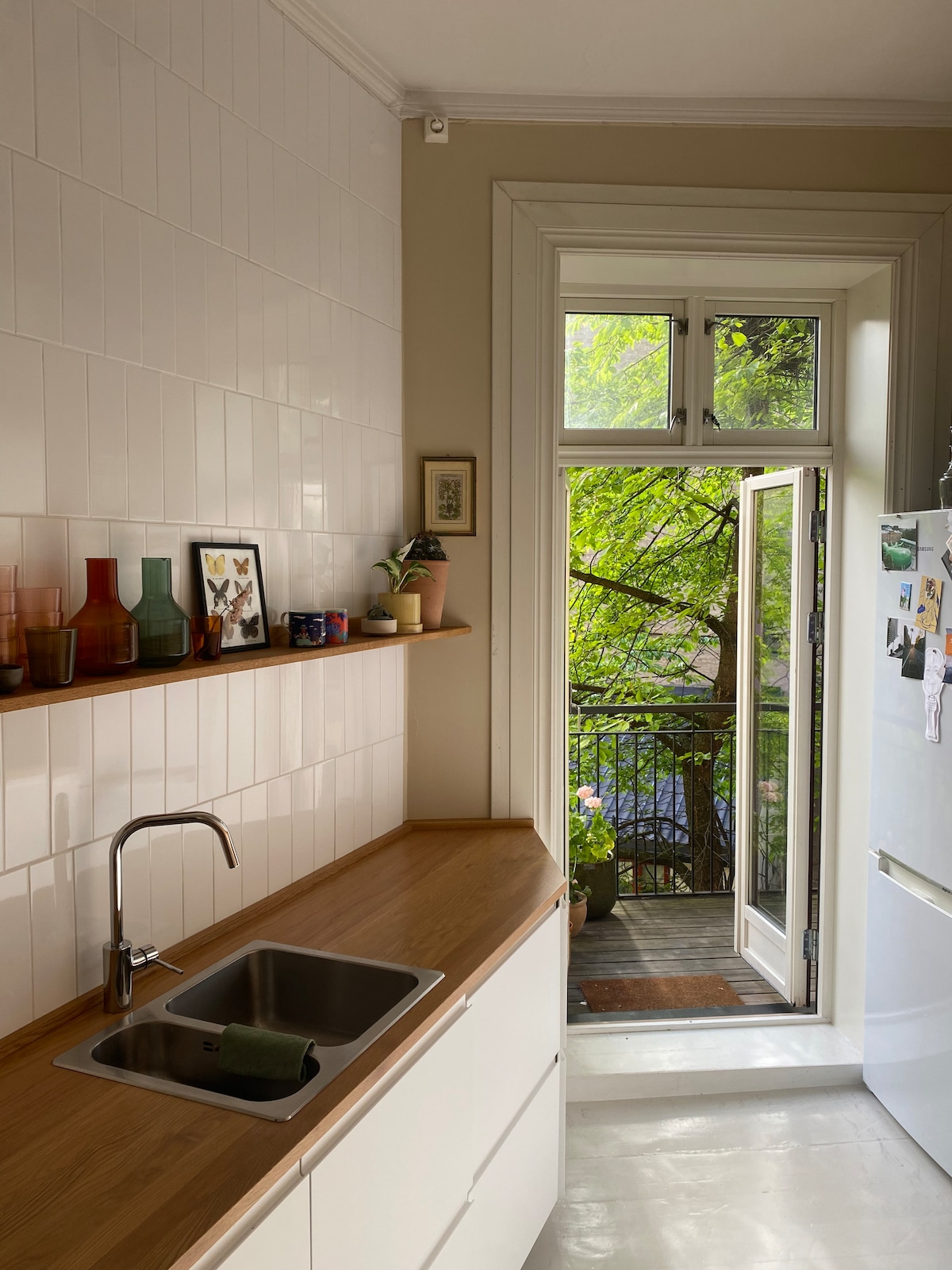 The kitchen features a sleek design with white cabinetry and a wooden countertop. A double sink is positioned near a window that opens to a balcony adorned with plants. Colorful glassware is displayed on a shelf above, enhancing the bright atmosphere.