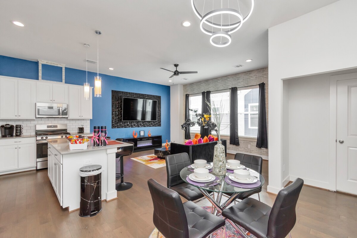 The open-concept living area combines a modern kitchen with a dining space. A sleek dining table is set for four, surrounded by black chairs. A comfortable seating area features a large TV mounted on the wall, with floor-to-ceiling windows dressed in dark curtains.