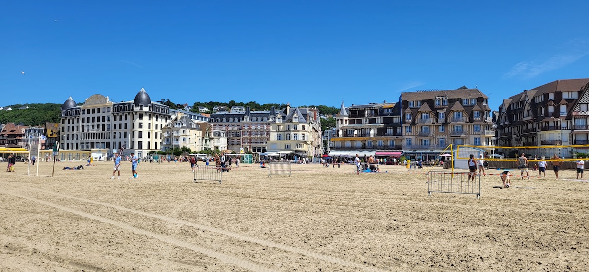 A sandy beach is filled with visitors enjoying the sunny weather. Volleyball nets are set up in the foreground, while a variety of buildings, showcasing diverse architectural styles, line the beach in the background. The clear blue sky enhances the inviting ambiance of the location.