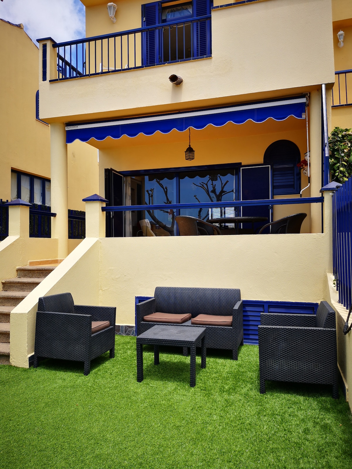 A welcoming outdoor seating area is presented, featuring a set of dark wicker furniture with cushions, arranged around a small table. The space is shaded by a blue awning and overlooks a manicured lawn, providing a peaceful ambiance for relaxation.
