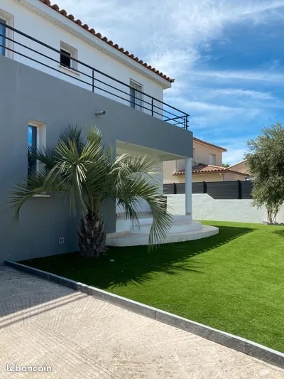 A modern home is set against a clear blue sky, featuring a neatly manicured lawn with a palm tree. The exterior includes a balcony with railings and a covered entrance area that provides shaded access to the front door.