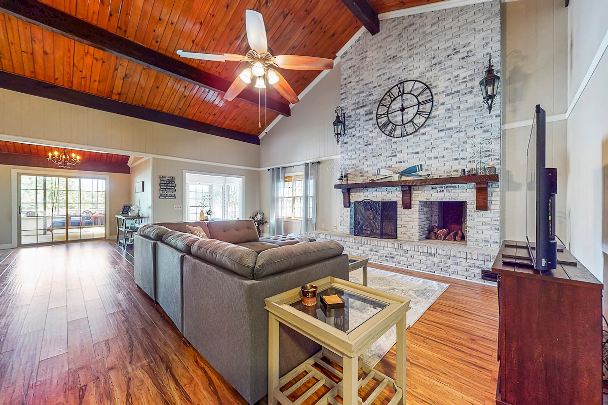 A spacious living room is highlighted by a large stone fireplace and a wooden beam ceiling. Comfortable seating is arranged with a coffee table in between. Natural light fills the room through large windows, offering a view of the outdoors.
