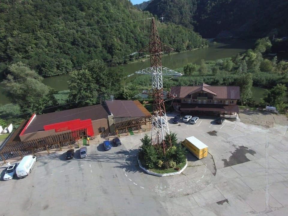 An aerial view captures a wide area featuring a rustic building with a red accent, surrounded by greenery. Adjacent vehicles are parked in a designated area, while a tall power tower stands nearby. A river is visible in the background, framed by lush hills.