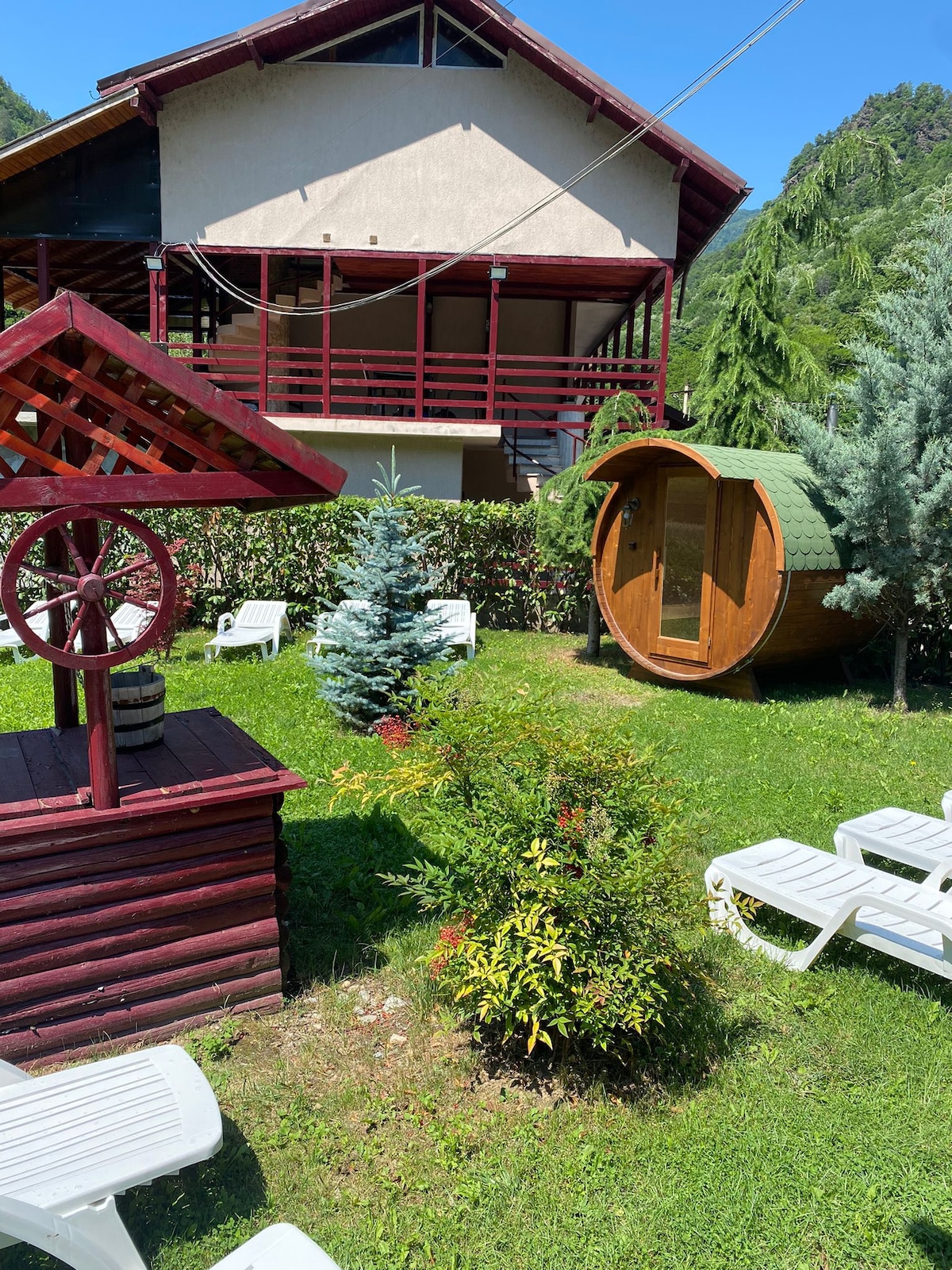 The image displays a spacious outdoor area featuring a well-maintained lawn. A wooden gazebo with a wheel design is visible in the foreground, alongside neatly arranged lounge chairs. A green sauna barrel is positioned nearby, surrounded by lush trees and shrubs, with a multi-level building in the background.