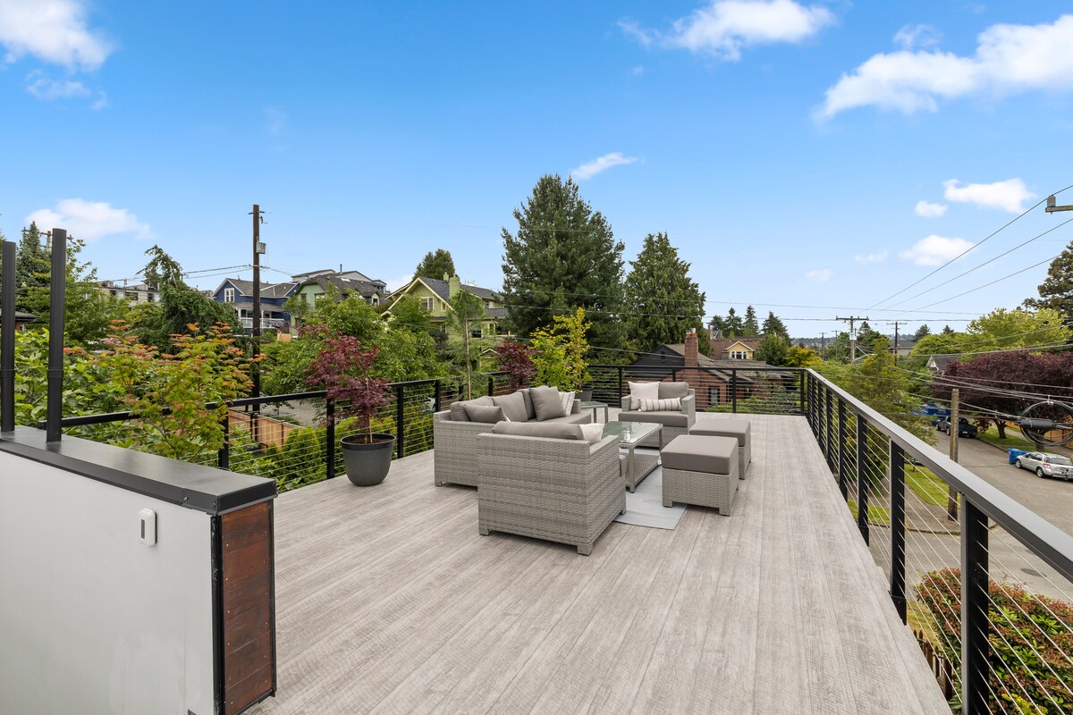 A spacious rooftop deck is presented, featuring comfortable seating arrangements around a modern coffee table. Surrounding greenery and distant residential views add to the serene atmosphere. The deck's wooden flooring complements the contemporary design, inviting relaxation in an outdoor setting.