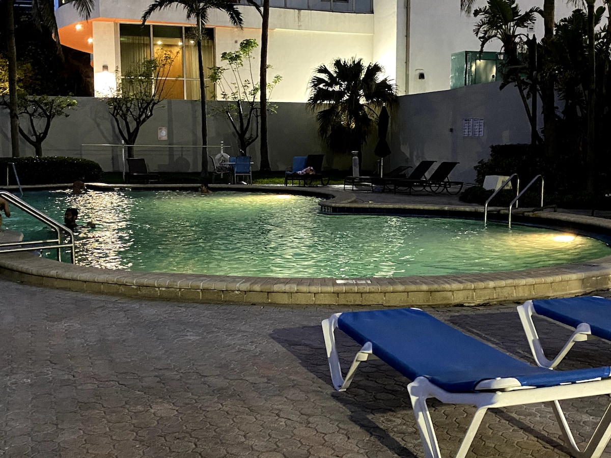 A heated swimming pool is surrounded by tropical landscaping, illuminated softly by lights at dusk. Several lounge chairs are positioned nearby, while palm trees frame the pool area. The pool's curved shape invites relaxation, reflecting the tranquil environment.