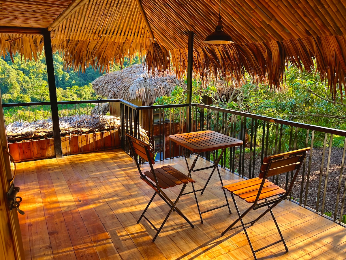 An outdoor deck is featured, framed by a thatched roof. Two wooden chairs are positioned around a small, rectangular table, offering a cozy space to enjoy the surrounding greenery. Sunlight casts warm tones on the wooden floor, enhancing the natural ambiance.