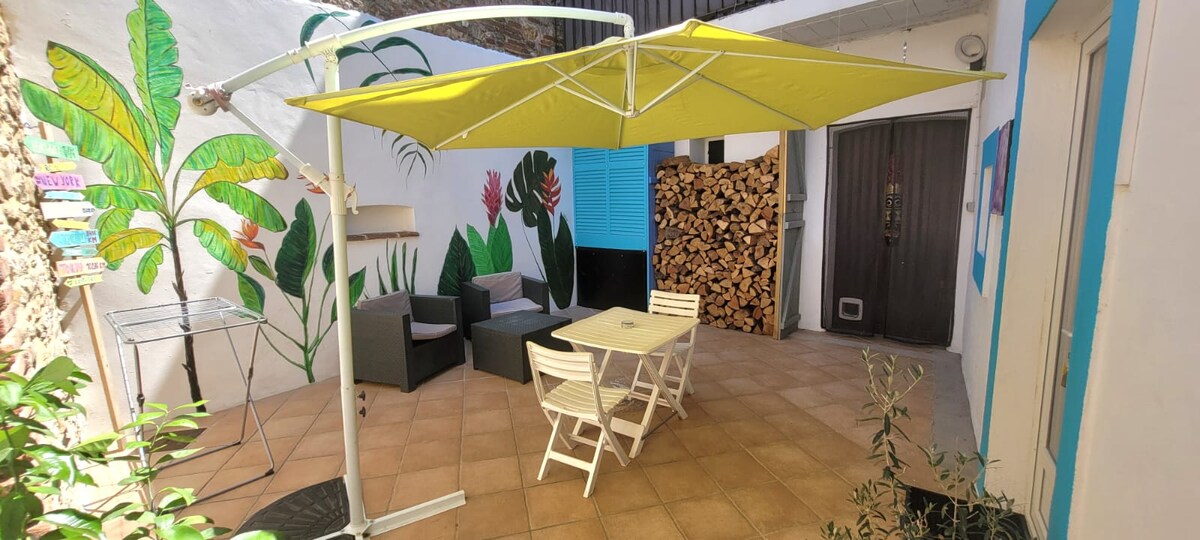 A private patio is presented, featuring a yellow canopy providing shade. A small table is surrounded by four white folding chairs, while two dark seating options are positioned against a vibrant mural of tropical plants. Stacked firewood is visible in the background.