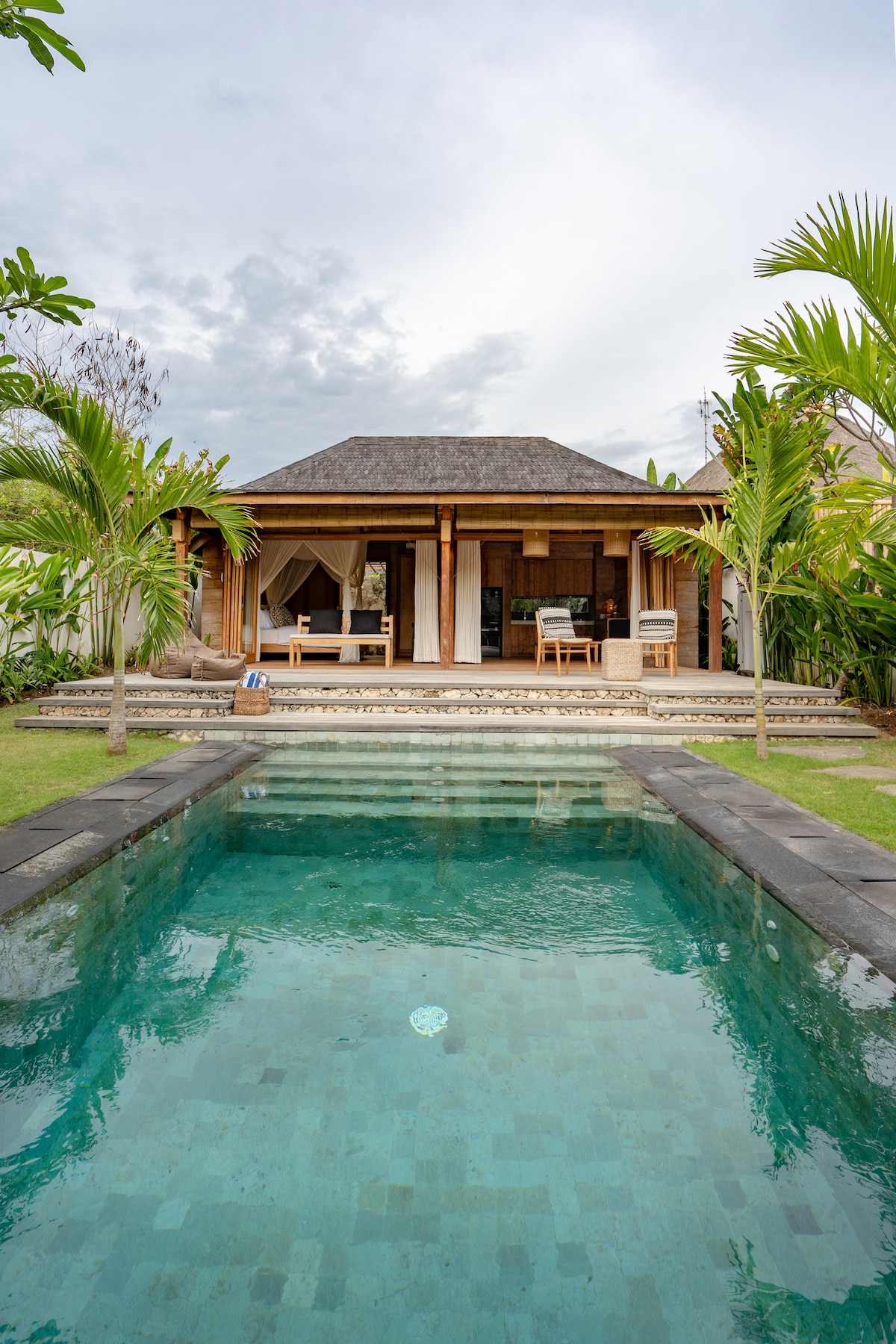 The villa is seen from the pool area, featuring a modern design with a thatched roof. A clear pool reflects the surrounding greenery, while two lounge chairs are positioned on the patio. Lush tropical plants frame the entrance to the villa.