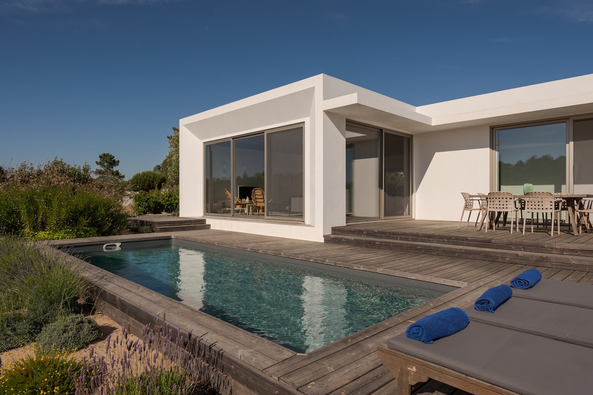 A contemporary house is shown with expansive glass windows offering views of the surrounding landscape. A wooden deck surrounds a rectangular pool, lined with lounge chairs and neatly rolled towels. Outdoor dining furniture is visible, allowing for relaxed al fresco meals.