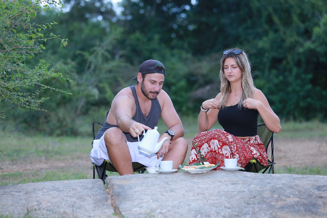 Two individuals are seated on portable chairs on a rocky surface, enjoying a meal. A white teapot and two cups are placed on a small plate. The background features greenery, suggesting a natural setting.